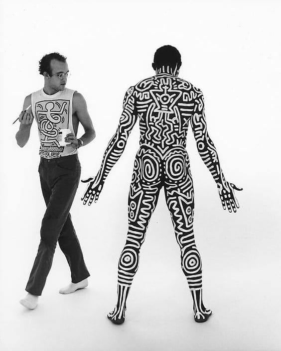 A white men with painting utensils and a black man with a painted body. 